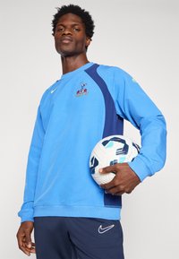 Zils džemperis ar tumši ziliem akcentiem un izšuvumu logotips; garās piedurknes un apaļa kakla izgriezums. Balts futbola bumbas turēšana vienā rokā.