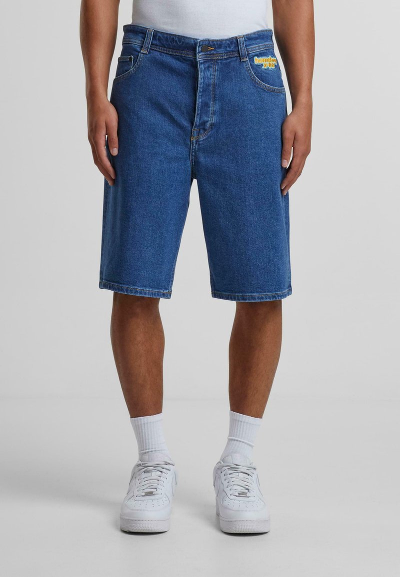 Pantalones cortos de denim en azul oscuro con corte recto, cinco bolsillos y logo de la marca en amarillo en la parte frontal. Llevados con zapatillas blancas y calcetines.