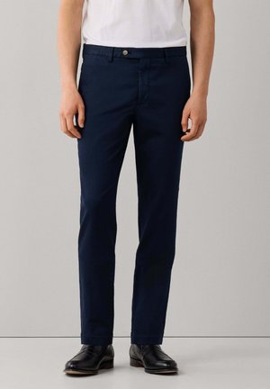 Man draagt marineblauwe slim-fit broek, wit overhemd en zwarte nette schoenen, staand tegen een effen lichte achtergrond.