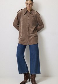 Brun kordfløjl skjorte med knapper og stort krave, parret med høj talje blå jeans og brune højhælede støvler i en neutral indstilling.