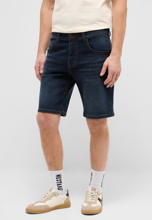 Mann trägt dunkelblaue Jeans-Shorts, weiße Socken mit der Aufschrift "MUSTANG", beige und weiße Sneakers und ein helles Hemd, steht vor einem grauen Hintergrund.