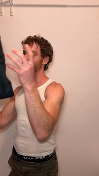 Homme en débardeur blanc et pantalon cargo tenant un vêtement noir devant un mur beige uni avec une tringle à rideau au-dessus.