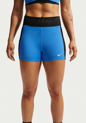 Atletyczna osoba nosząca niebieskie sportowe spodenki Nike z czarnym pasem i bocznymi panelami, stoi z rękami swobodnie opuszczonymi po bokach.