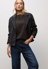 Dunkelgrau, maßgeschneiderte Jacke mit Reverskragen über einem braunen Pullover, kombiniert mit dunkelblauen Jeans. Glattes Material mit strukturierter Passform.
