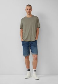 T-shirt vert olive clair, à manches courtes, coupe décontractée. Short en denim bleu foncé, avec les ourlets retroussés. Baskets blanches avec des chaussettes rayées noires et blanches.