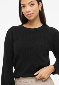 Mustast soonikkoesist kampsunist, millel on pikad varrukad ja ümara kaelusega, pehme tekstuuriga ja lõdva lõikega. Ühitatud beežide pükstega.