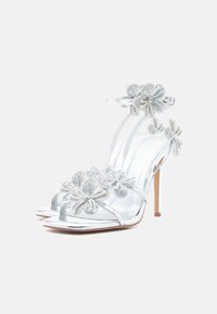 Sidabrinės aukštakulnės sandalai turi skaidrias dirželių detales, puoštas karoliukų gėlių ornamentais, ir uždarą kulkšnies dirželį gale.