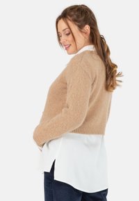 Beige cropped trui van zacht, pluizig materiaal, gelaagd over een wit overhemd met knopen. Heeft lange mouwen en een geribbelde onderrand.