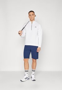 Pullover atletico bianco con zip e righe testurizzate, abbinato a pantaloni corti blu. Il modello indossa sneakers bianche con dettagli neri e calze a righe.