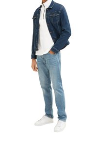 Chaqueta de mezclilla en azul oscuro, polo blanco con cremallera, jeans azul claro y zapatillas blancas. La chaqueta cuenta con bolsillos y un cierre de botones.