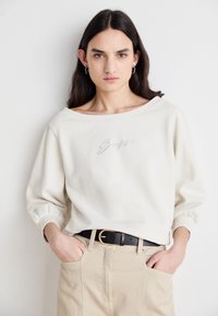 Licht beige oversized sweatshirt met een wijde halslijn en gathered mouwen, met subtiele strassteentjes borduursel dat "Boss" spelt. Gecombineerd met hoge taille beige broek en een zwarte riem.