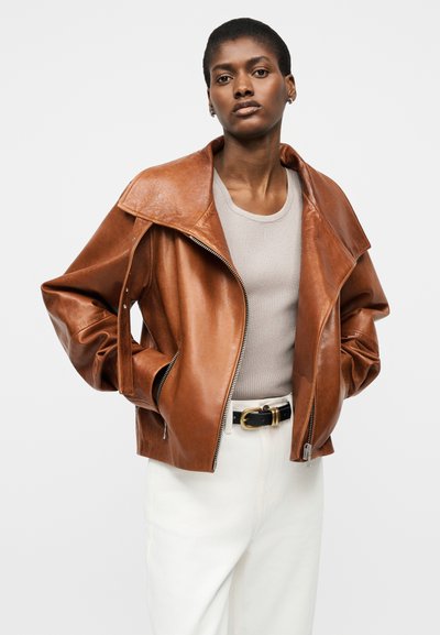 Γυναίκα που φοράει καφέ δερμάτινο μπουφάν πάνω από ανοιχτό μπεζ τοπ και άσπρο παντελόνι με μαύρη ζώνη, στέκεται με τα χέρια στις τσέπες του μπουφάν.