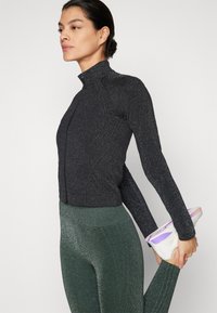 Sort zip-up jakke med høj krave, der har et skinnende mønster. Grønne, tætsiddende leggings med en subtil glans. Hvide sportsko synlige.