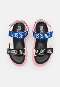 Musta pohjallinen sandaalit, joissa on vaaleanpunaiset reunat, siniset ja mustat hihnat, joihin on merkitty "Moschino", valkoista taustaa vasten.