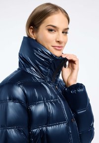 Marineblauwe gewatteerde jas met een hoge kraag, glanzende textuur, horizontaal gestikt patroon en ritssluiting. Heeft een snug fit rond de polsen.