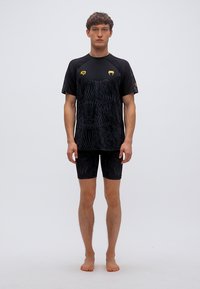 Μαύρο αθλητικό μπλουζάκι με υφασμάτινο σχέδιο, κοντά μανίκια και χρυσές λεπτομέρειες· συνδυάζεται με μαύρο εφαρμοστό σορτς συμπίεσης, και τα δύο στενά.