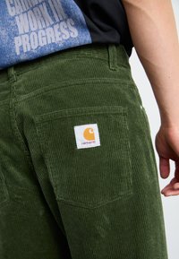 Zelene kordeljske hlače z izrazitimi vertikalnimi rebri, z zadnjim žepom, ki ima oznako Carhartt v oranžni in beli barvi.