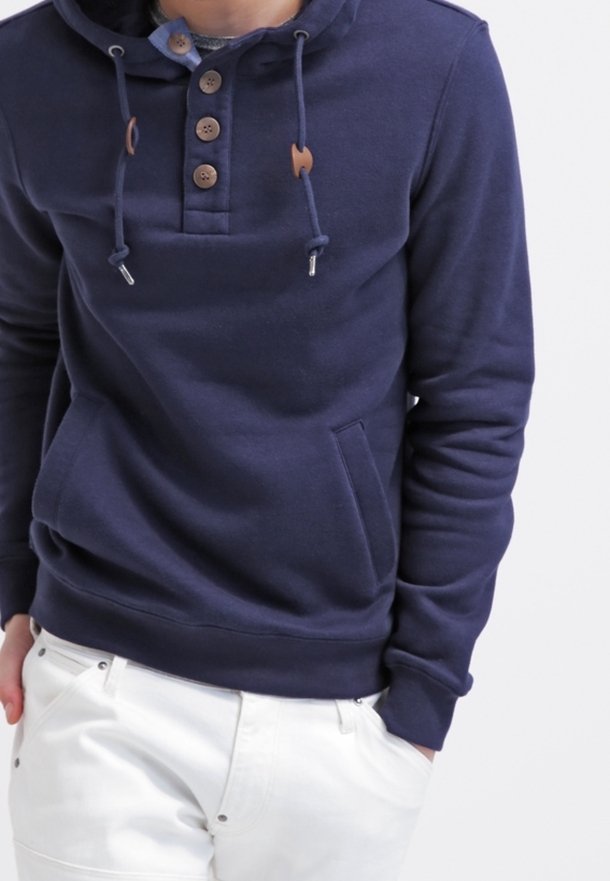 Osoba ubrana w granatową bluzę z kapturem z drewnianymi guzikami i białe spodnie, z rękami w kieszeniach, na jednolitym tle.