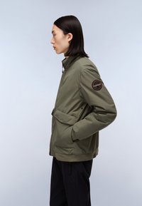 Olijfgroene jas met een staande kraag, voorrits, zijzakken en een zwarte patch op de linker mouw. Glad textuur en modern ontwerp.