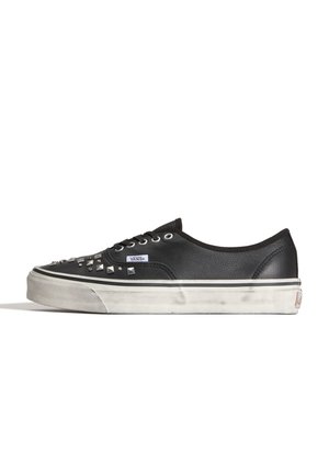 Zapatilla baja negra Vans con tachuelas metálicas en la puntera, suela intermedia blanca con textura desgastada y cordones negros sobre fondo blanco.