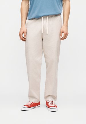 Muž v béžových nohaviciach so sťahovacou šnúrkou, modrej košeli a červených nízkych teniskách stojaci pred jednoduchým svetlým pozadím.