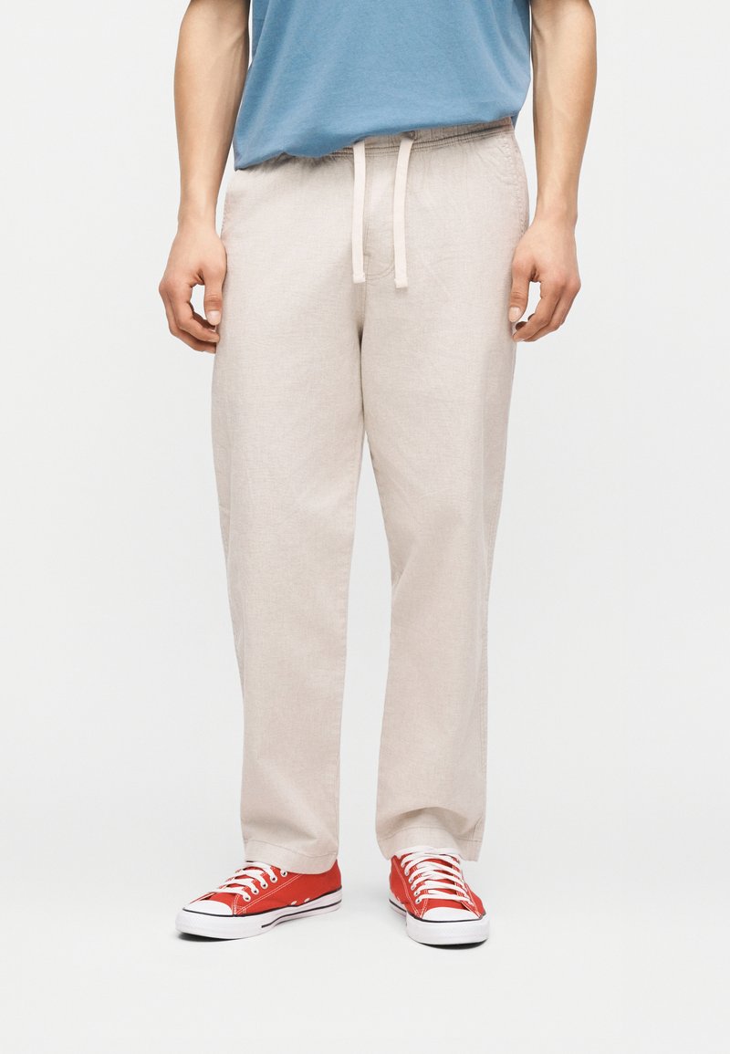 Άνδρας που φοράει μπεζ παντελόνι με κορδόνι, μπλε πουκάμισο και κόκκινα αθλητικά παπούτσια χαμηλής κοπής, στέκεται μπροστά σε ένα απλό φωτεινό φόντο.