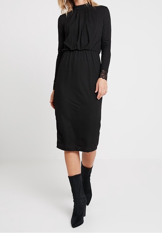 Abito nero a coste con maniche lunghe, dettaglio arricciato sul fronte e inserti in pizzo ai polsini. Vita aderente, design fino al ginocchio, abbinato a stivali neri.