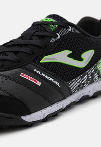 Pantof de sport negru cu partea superioară din plasă, accente verzi, logo gri și bază sintetică texturată. Are o talpă cu model alb și negru.