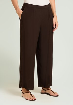Femme portant un pantalon ample marron foncé avec des poches latérales et des sandales plates marron, debout sur un fond gris clair.