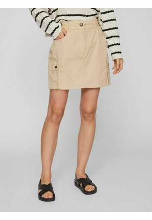 Falda de algodón beige con dos bolsillos laterales, cierre de botón y dobladillo recto. Llevada con sandalias negras de tiras cruzadas.