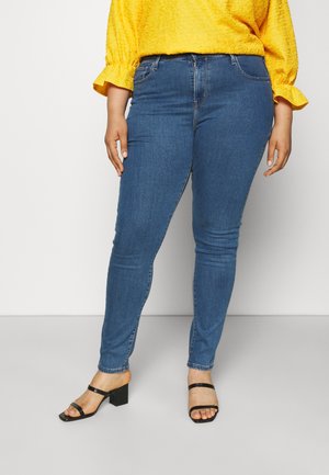 Femme portant un jean skinny bleu, un haut texturé jaune avec des manches à volants, et des sandales noires à petits talons devant un fond uni.