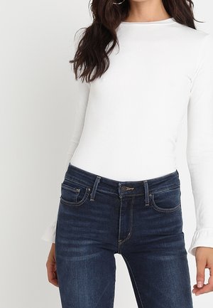 Mujer con una camiseta blanca de manga larga ajustada y jeans de talle alto azul oscuro, con cabello ondulado oscuro y pendientes de aro.