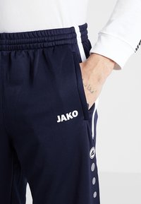 Osoba v tmavomodrých sportovních kalhotách JAKO s bílými pruhy a logy, ruka vložená v pravé kapse, viditelné tetování na zápěstí.