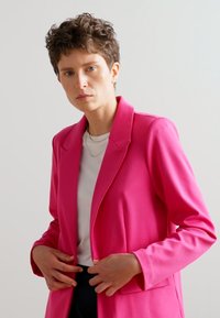 Skreddersydd rosa blazer med en glatt tekstur, klaffelapper og en enkelt knappelukking, båret over en hvit skjorte og kombinert med mørke bukser.