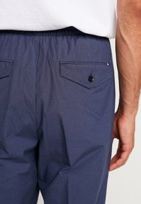 Navyblå byxor med elastisk midja och knäppta bakfickor, bära av en person i vit skjorta med synlig högerarm.