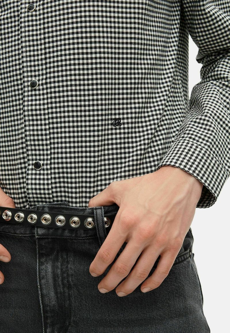 Schwarz-weiß kariertes Hemd mit einer Vordertasche, kombiniert mit einem dunklen Denim-Gürtel mit metallischen Druckknöpfen und einer strukturierten Oberfläche.