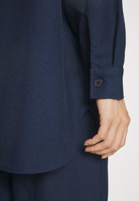 Mörkblå skräddarsydd skjorta med en mjuk textur, med en knäppt manschett och en något rundad linning. Närbild på ärm och hand.