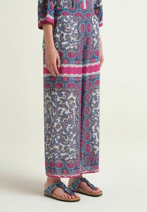 Pantaloni a gamba larga con motivo paisley rosa, blu e bianco, abbinati a sandali piatti blu con perline su uno sfondo chiaro.