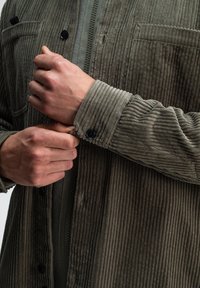 Olivegrüner Cordjacke mit vertikalen Rippungen, einem Reißverschluss vorne und schwarzen Knopfakzenten; verfügt über eine Brusttasche und strukturierte Ärmelbündchen.