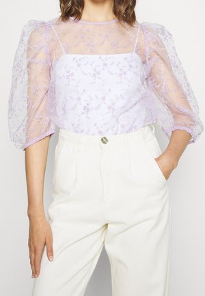 Kobieta ubrana w przezroczystą lawendową bluzkę z haftowanymi kwiatami na bufiastych rękawach oraz wysokie, białe spodnie, z jedną ręką w kieszeni.