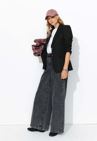 Blazer noir à rayures sur un t-shirt blanc, jean large gris, escarpins noirs studdés et une casquette rose poussiéreux. Tenant une écharpe à motif rouge.