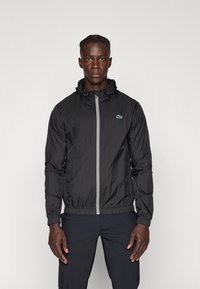 Zwarte lichtgewicht jack met rits en capuchon, met elastische manchetten en zoom, featuring een klein groen logo op de borst en een gladde, iets glanzende textuur.