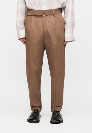 Άνδρας που φορά καφέ πλισέ παντελόνι με ψηλή μέση και ζώνη, λευκό μακρυμάνικο πουκάμισο μέσα στη μέση και μαύρα δερμάτινα σανδάλια με ανοιχτά δάχτυλα.