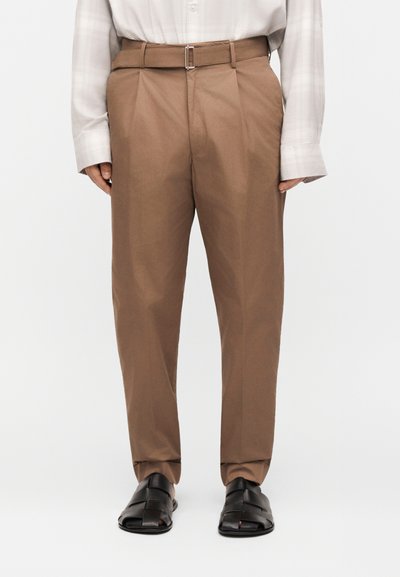 Άνδρας που φορά καφέ πλισέ παντελόνι με ψηλή μέση και ζώνη, λευκό μακρυμάνικο πουκάμισο μέσα στη μέση και μαύρα δερμάτινα σανδάλια με ανοιχτά δάχτυλα.