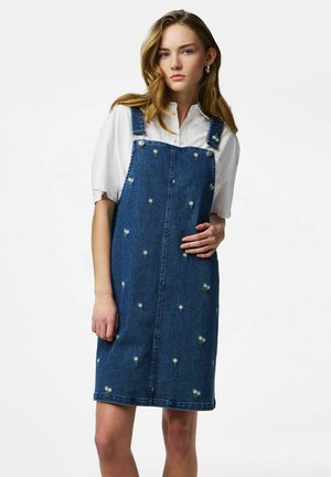 Ung kvinne med bølgete hår som har på seg en hvit kortermet skjorte under en blå denimkjole brodert med små hvite blomster.