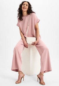 Helles pinkes Outfit bestehend aus einem lockeren Oberteil und weit geschnittenen Hosen. Dazu eine cremefarbene Umhängetasche und beigefarbene Absatzsandalen. Glatte Textur.