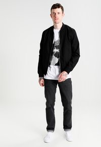 Sort bomberjakke over en hvid T-shirt med grafisk print, parret med sorte jeans og hvide sneakers.