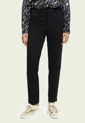 Personne portant un pantalon noir droit, une chemise à manches longues à motif rentrée, et des baskets beige et blanches avec les lacets non attachés.