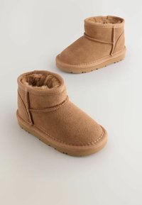 Botine din suede bej cu o căptușeală din blană moale, talpă din cauciuc plat și accente de cusătură. Au un design cu guler pentru o ajustare strânsă.