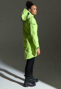 Limoengroene parka met een capuchon, voorzien van voorzakken en verstelbare manchetten, gecombineerd met zwarte slim-fit broek en stevige zwarte laarzen.